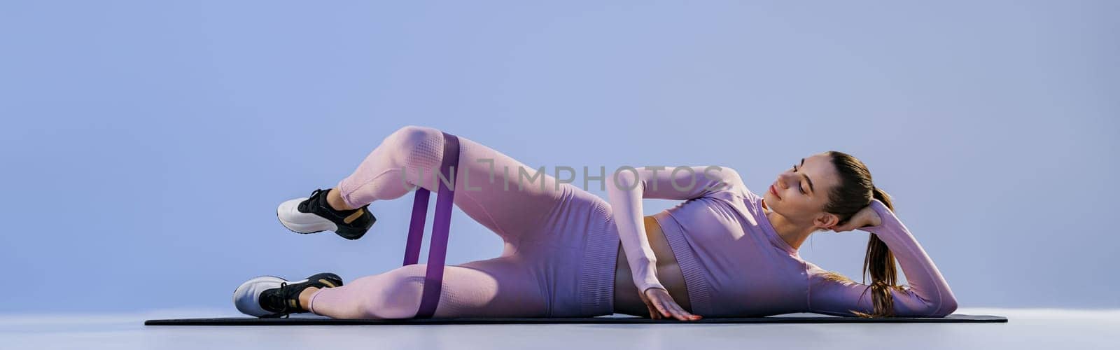 Sporty woman lying on yoga mat and trains legs with fitness elastic band on studio background by Yaroslav_astakhov