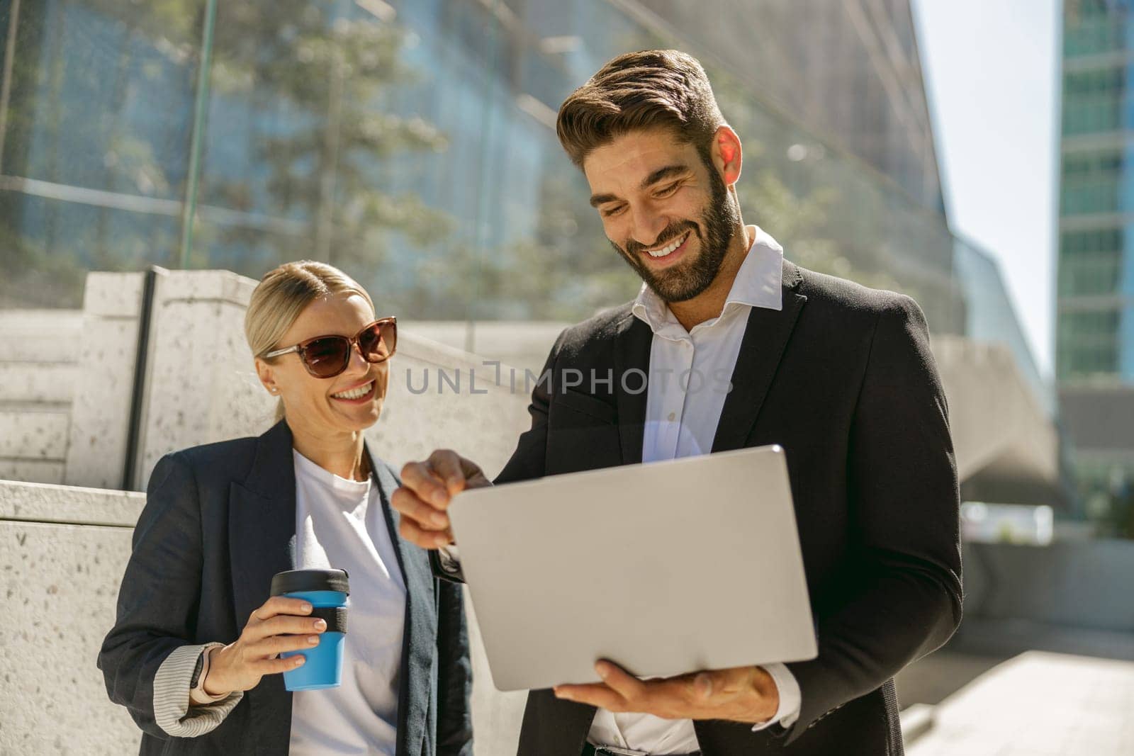 Man and woman in classic suit discussing business details and using laptop while standing outdoors by Yaroslav_astakhov