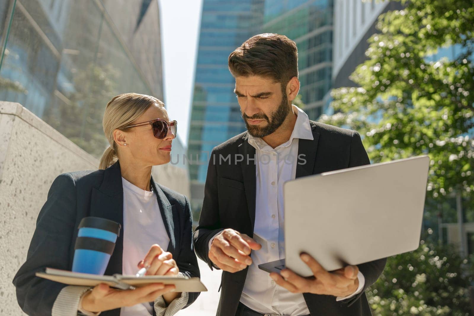 Man and woman in classic suit discussing business details and using laptop while standing outdoors