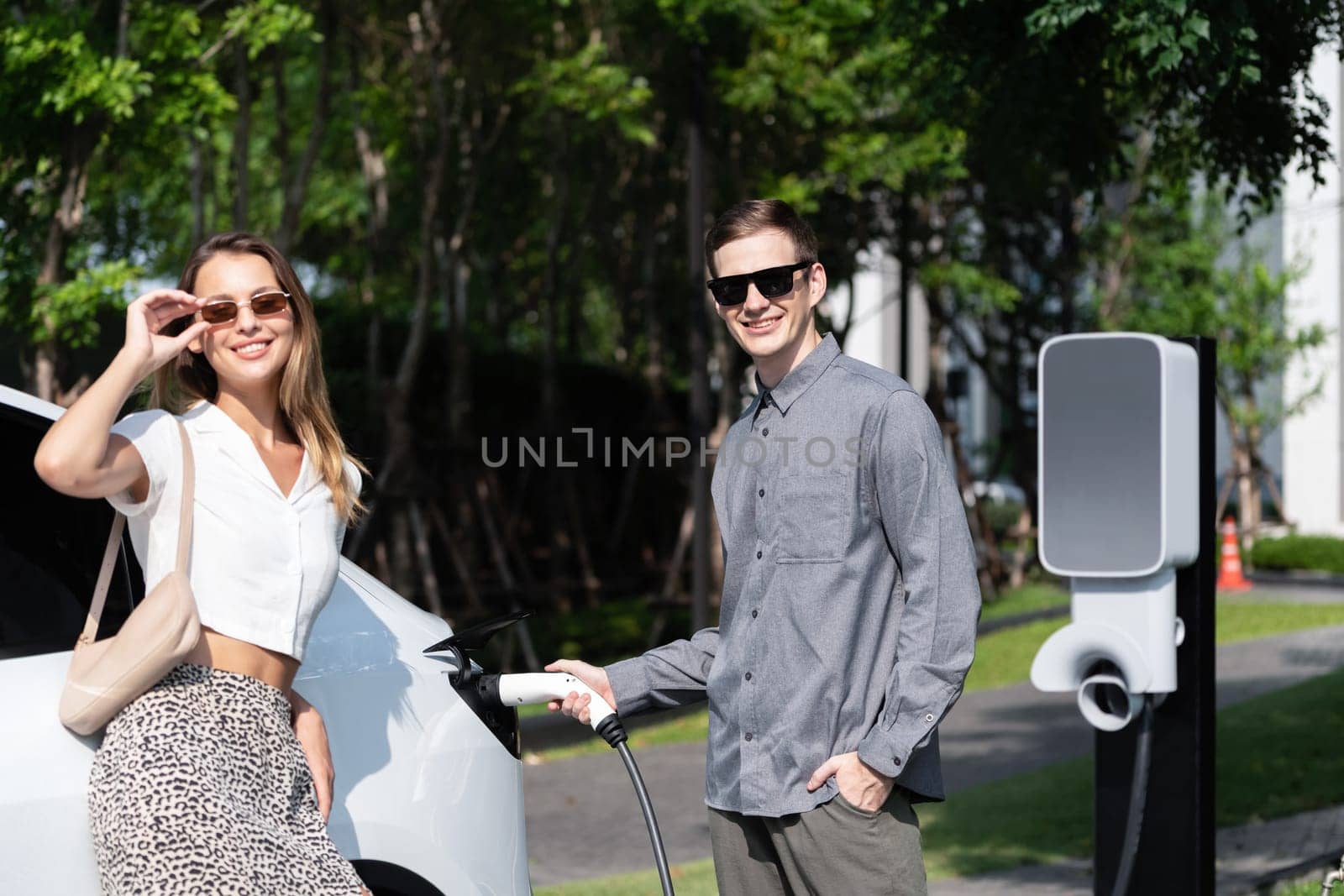 Young couple travel with EV electric car charging in green sustainable city outdoor garden in summer shows urban sustainability lifestyle by green clean rechargeable energy of electric vehicle innards