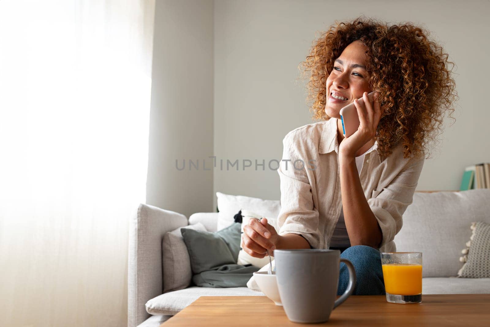 Young multiracial hispanic woman talking on the phone while having breakfast sitting on the sofa at home cozy living room. Copy space. Coffee and technology concept.