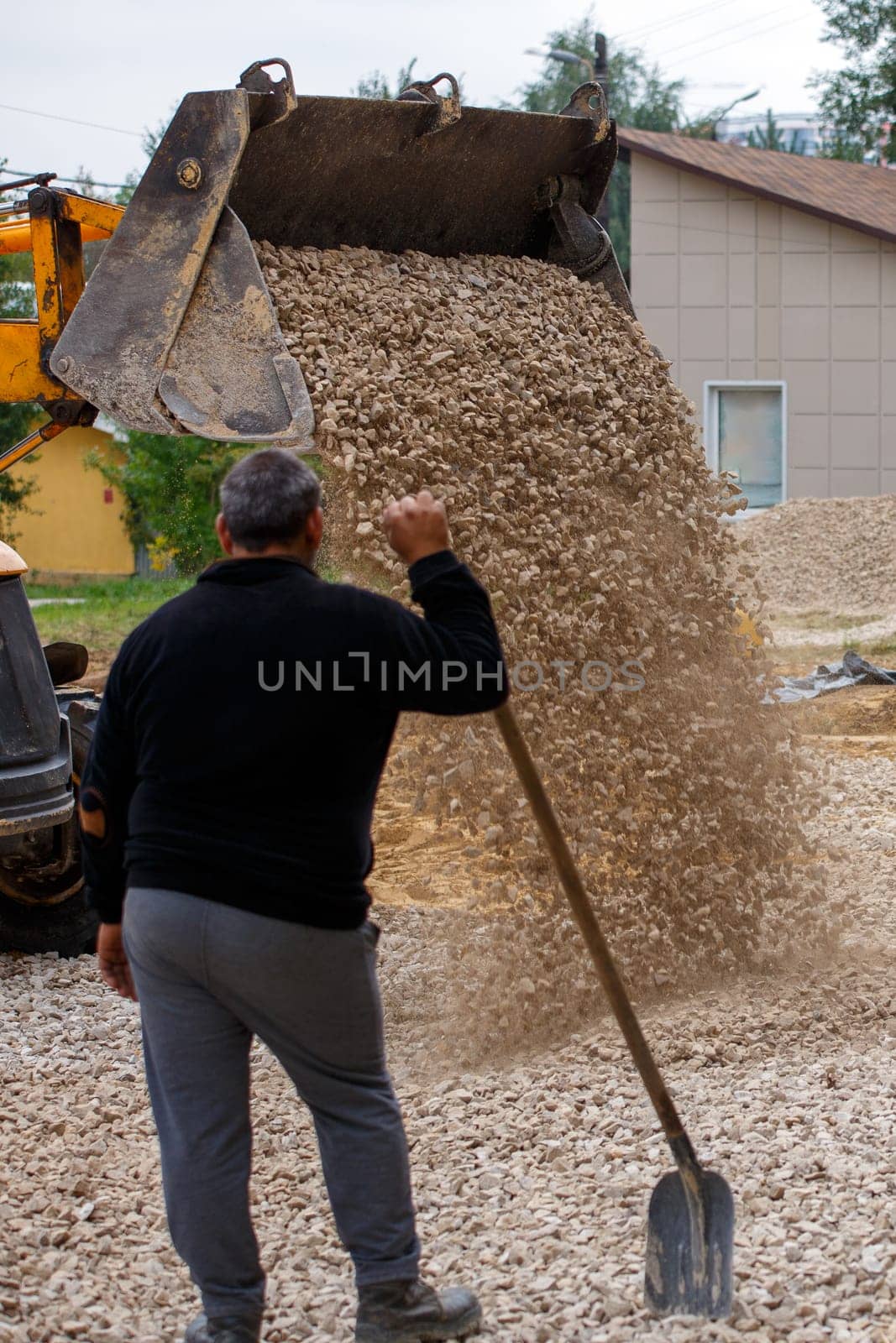 a bulldozer pours macadam from a bucket onto the ground at a construction site in front of gray-haired worker with shovel.