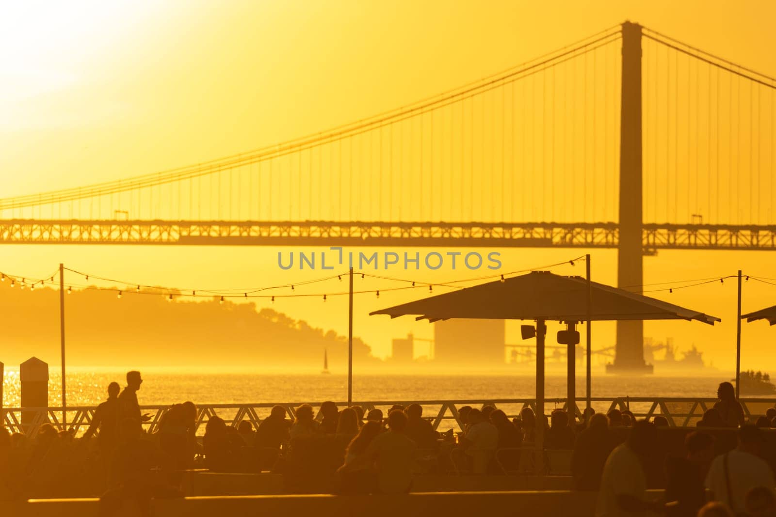 A Serene Evening by the Bridge: People Relaxing Under Umbrellas as the Ocean Glows with Sunset by Studia72