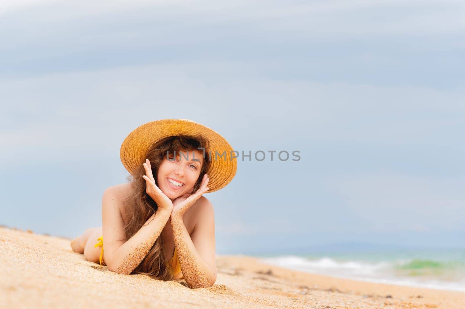 a woman in a bikini and a straw hat lies on a tropical beach. Beautiful girl lying and smiling on the sand while sunbathing during vacation