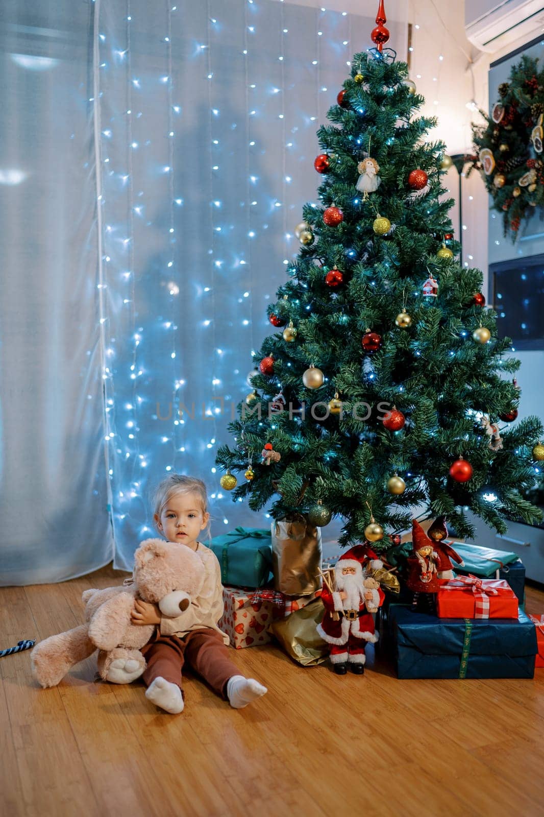 Little girl with a teddy bear sits on the floor next to gifts under the Christmas tree. High quality photo