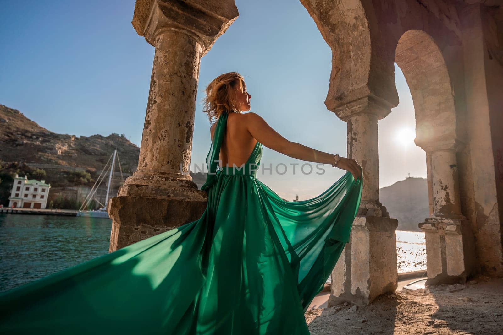 Woman dress sea columns. Rear view of a happy blonde woman in a long mint dress posing against the backdrop of the sea in an old building with columns