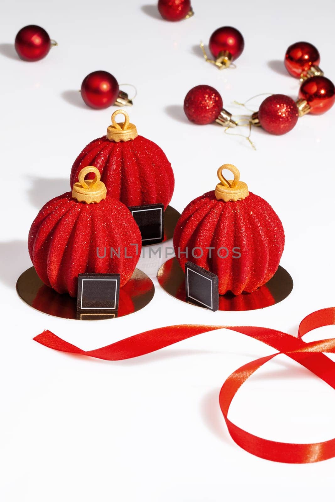 Three delicious pastries of chocolate mousse, brownie and cranberries in shape of Christmas balls covered with red and gold sugar glaze on table with ribbon. Festive collection of authors desserts