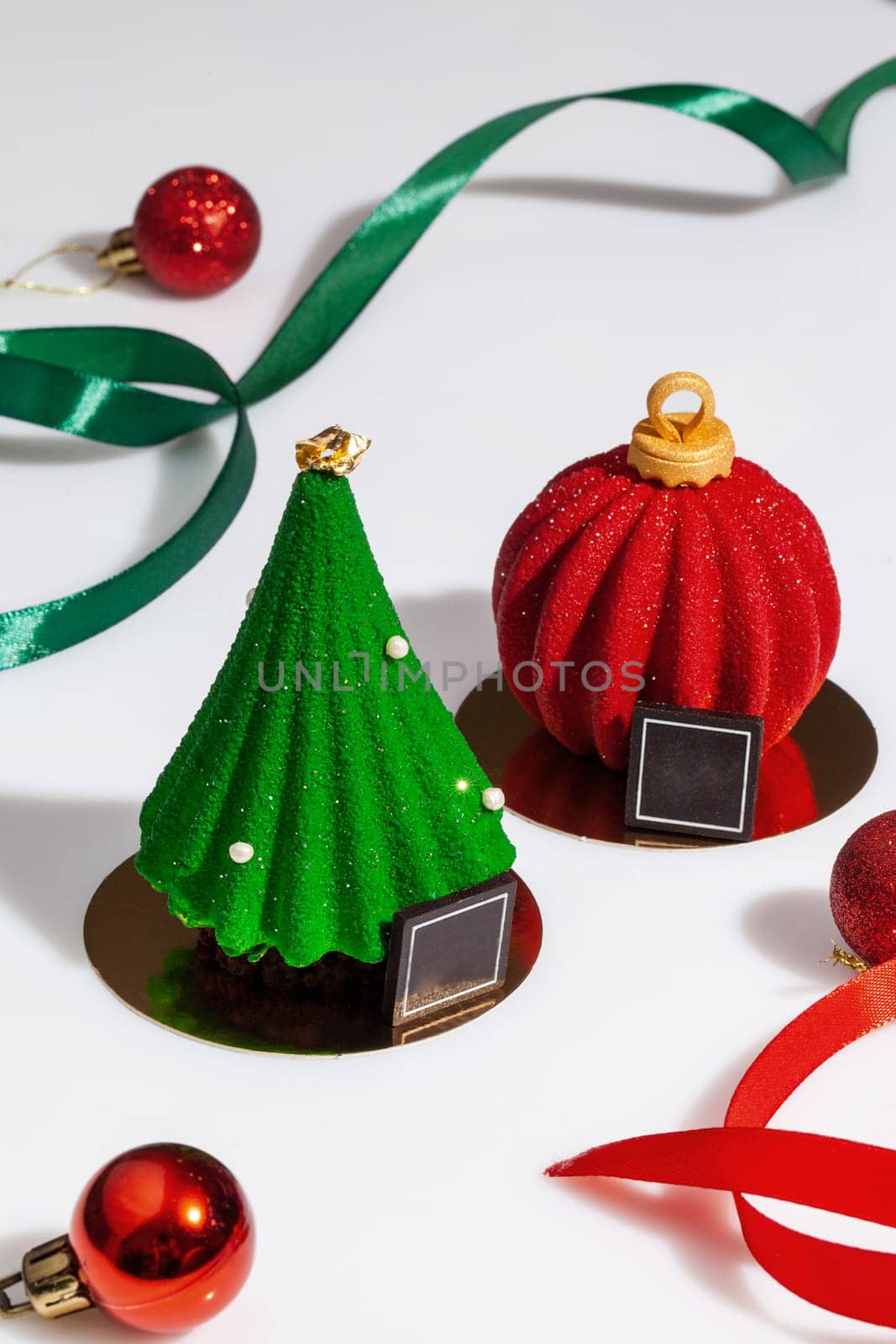 Holiday desserts in shape of green glazed fir and red ball surrounded by ribbons and Christmas tree toys on white background. Delicious treats for New Year celebration. Creating festive mood concept