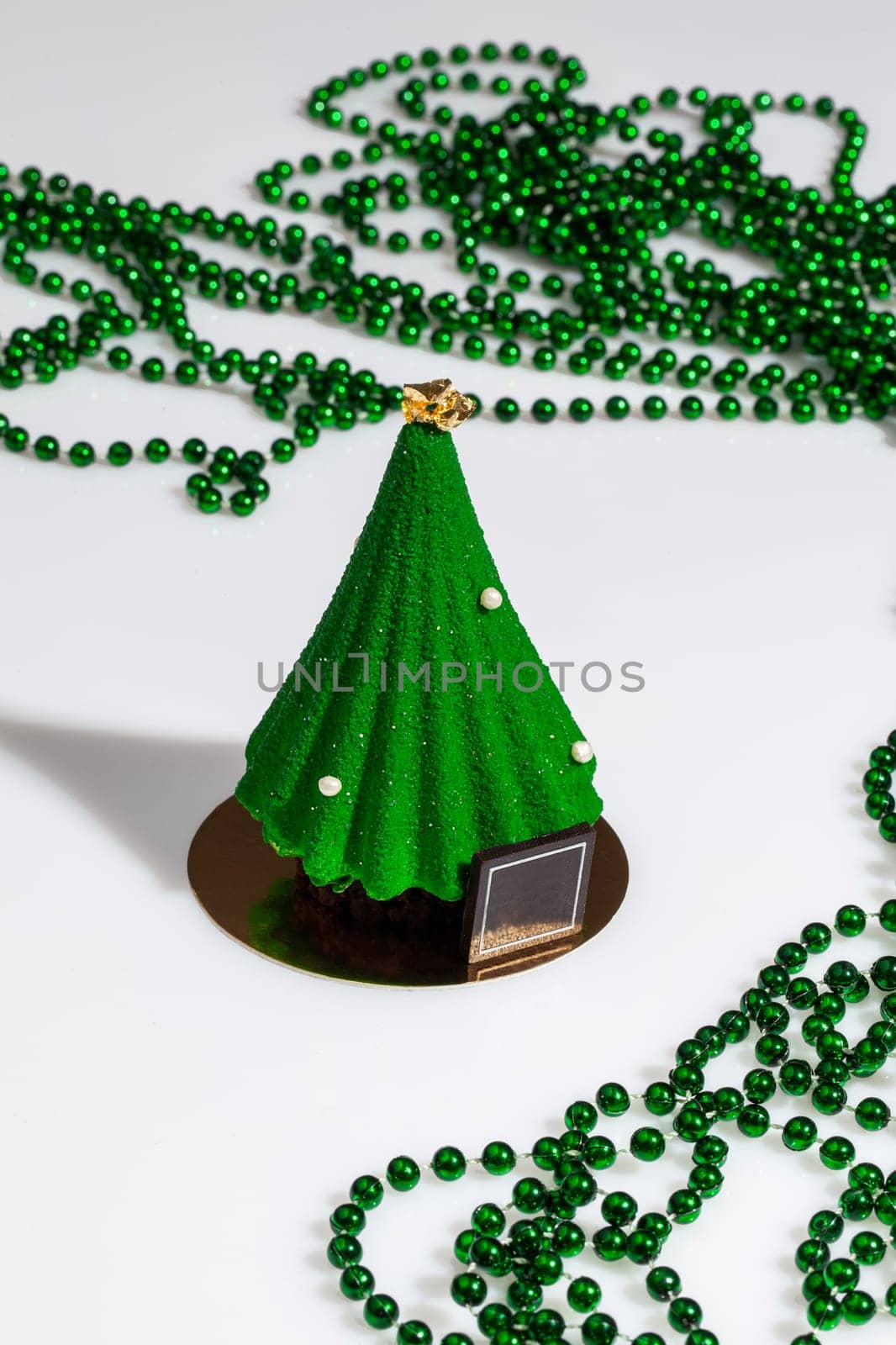 Christmas tree shaped pastry covered with scabrous green icing with golden topper and sugar drops on white background with strings of green beads. Festive decor and treats. Handmade authors dessert