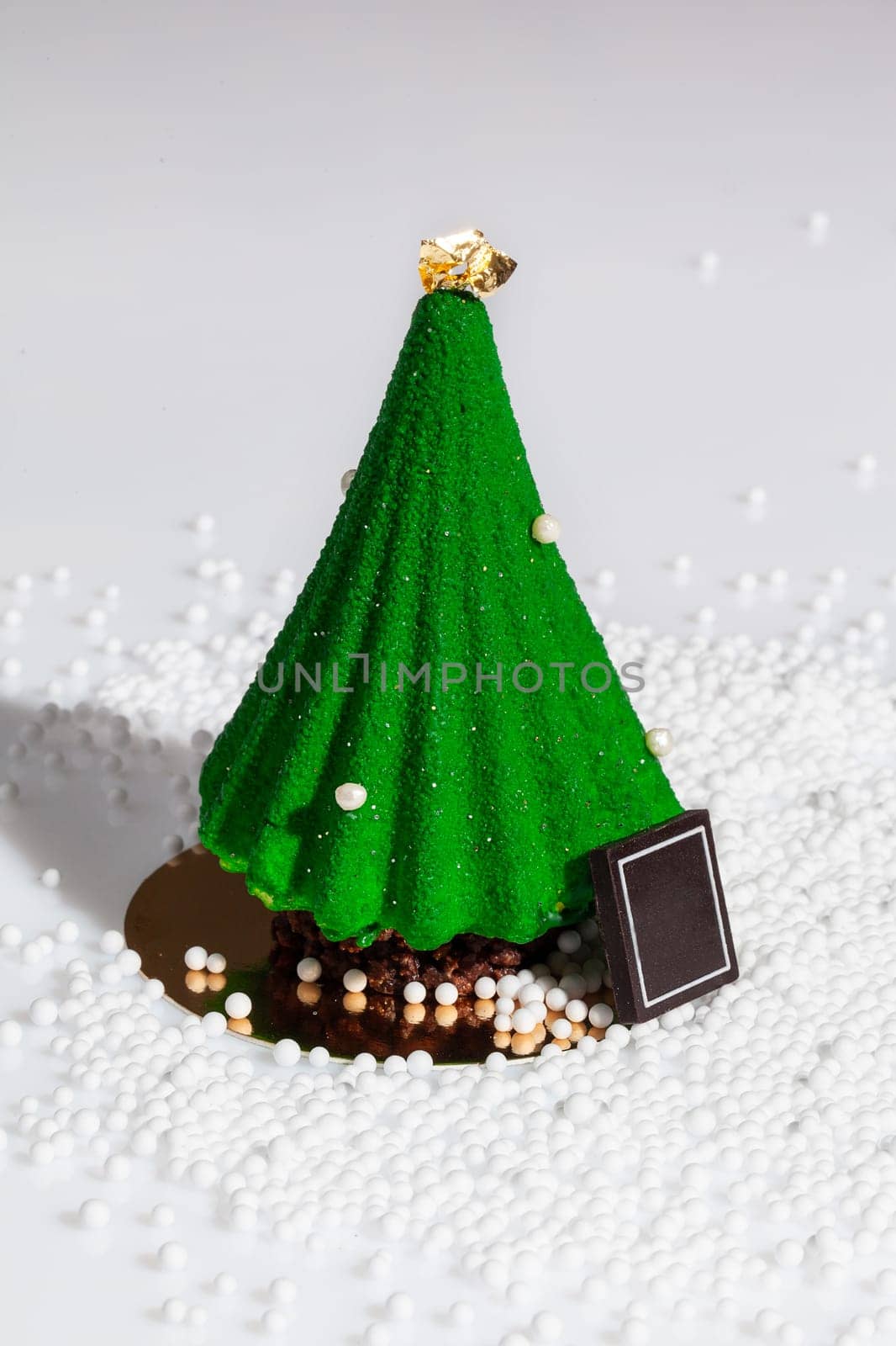 Delicious treats for New Year celebration. Handmade authors dessert in shape of green glazed Christmas tree with golden topper sprinkled with sugar beads on white background. Festive mood concept