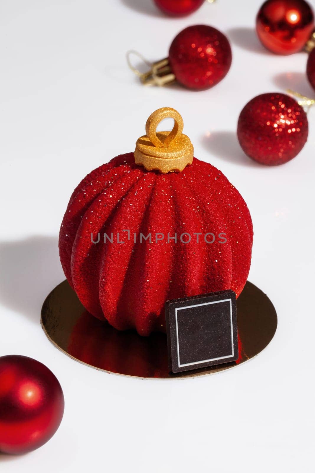 Red sparkling Christmas tree toys and ball shaped cake on white surface. Festive handmade dessert of chocolate mousse, cherry compote, hazelnut and almond biscuit covered with colored sugar icing