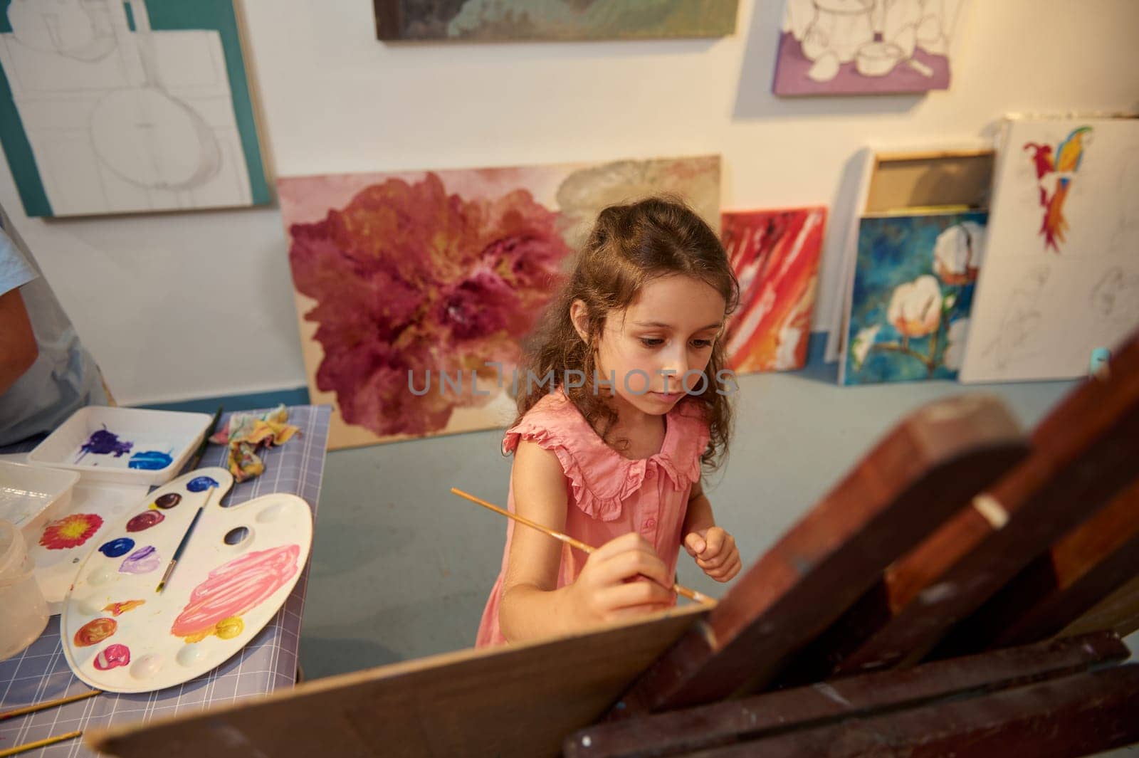 Adorable little girl drawing picture on canvas, standing by a wooden easel in creative art workshop. by artgf