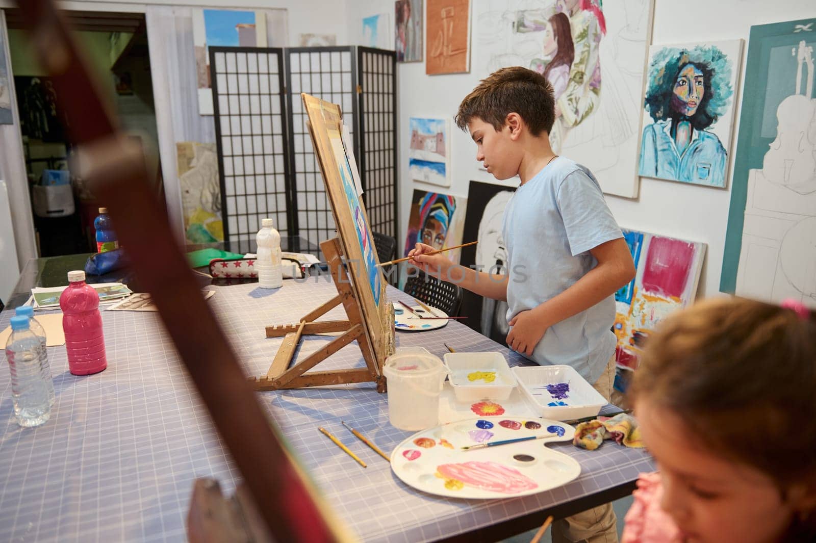 Talented elementary age kids, confident focused teen boy and girl, inspired artists painters standing by wooden easel and painting with watercolors on canvas, learning art in a cozy creative workshop