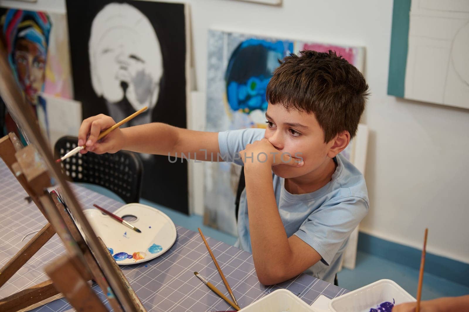 Teen boy expressing himself while drawing on canvas, holding paintbrush and oil painting pictures in creative workshop by artgf