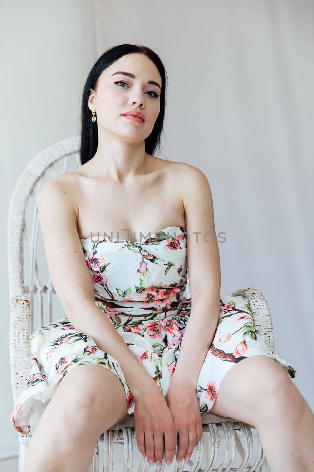 a beautiful woman in a floral dress sits in a chair in a bright room by Simakov