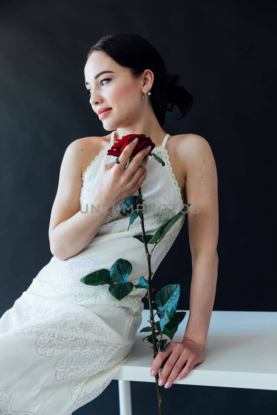 beautiful woman in a white dress sits with a red rose on a dark background
