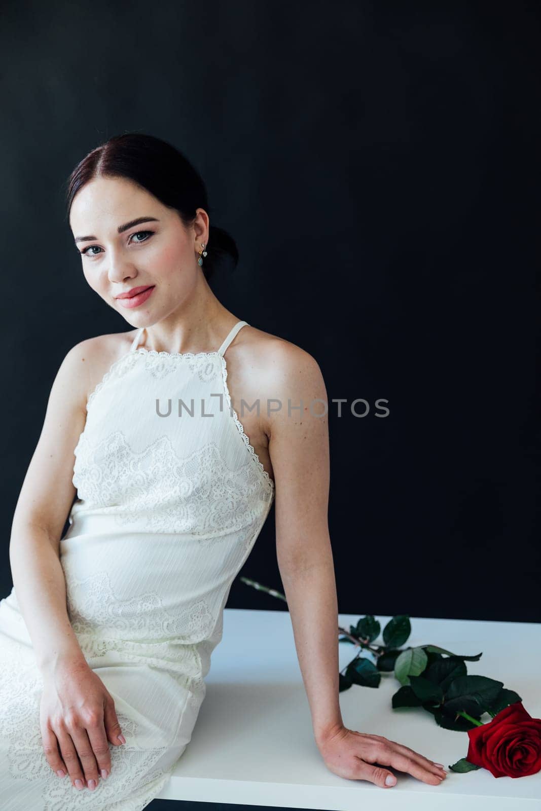 a beautiful woman in a white dress sits with a red rose on a dark background by Simakov