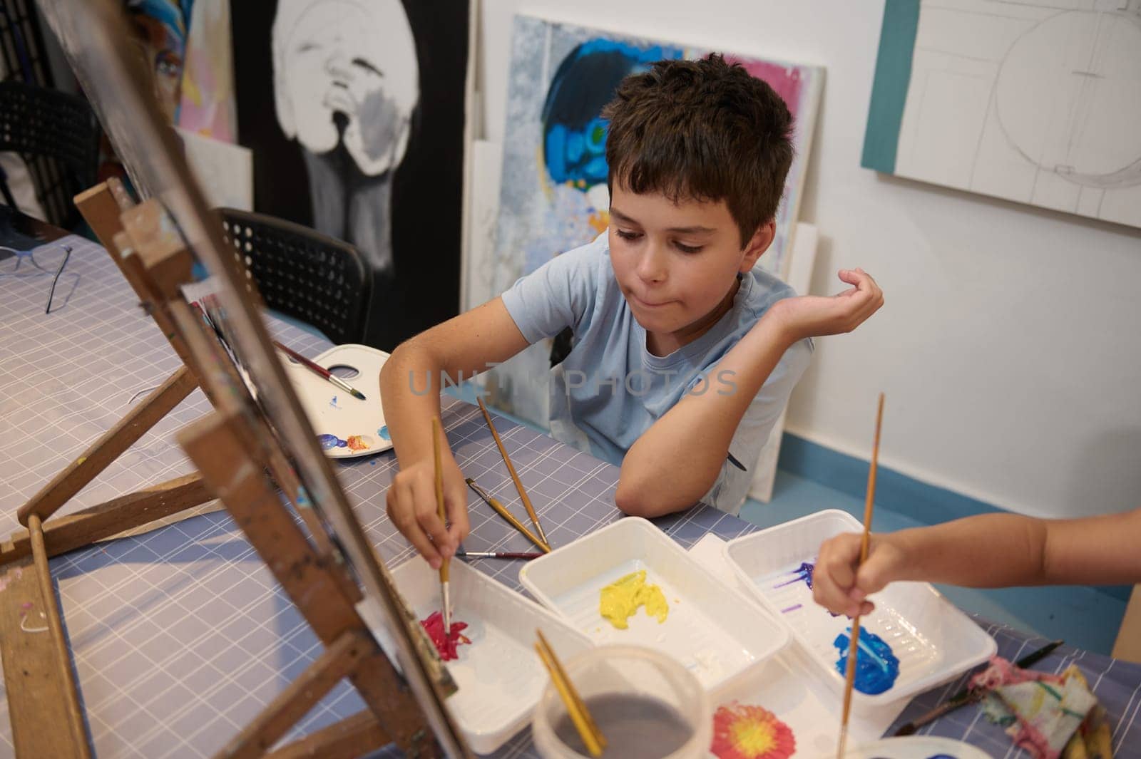 Authentic confident happy teenage boy, elementary age school student paints picture during creative art and craft class by artgf