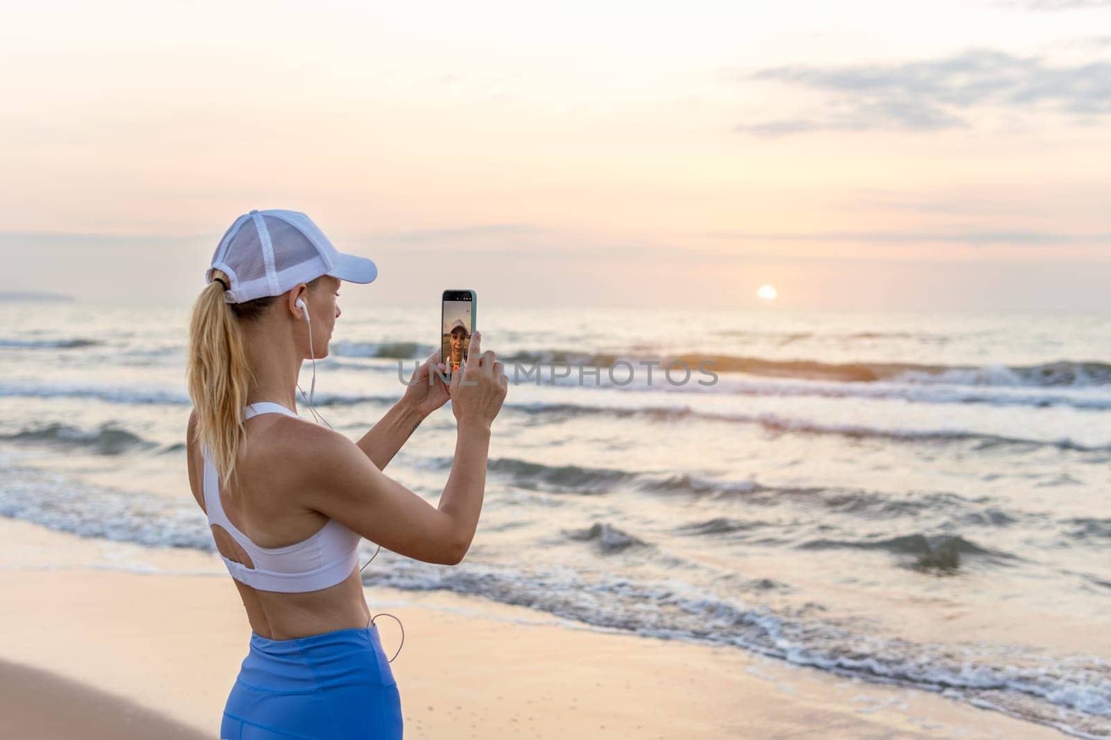 girl runner runs along the beach in sportswear. the girl takes a photo of the sunrise at sea by PopOff