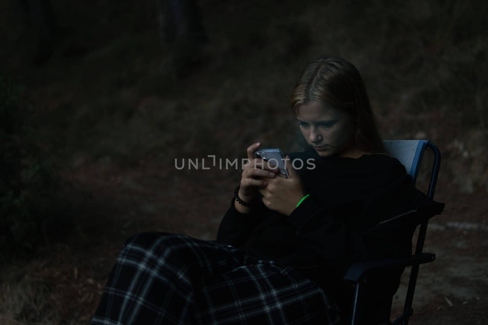 a teenage girl in casual clothes sits on a chair in a Camping and looks at the phone, a girl sits in the forest among the trees on a folding chair. High quality photo