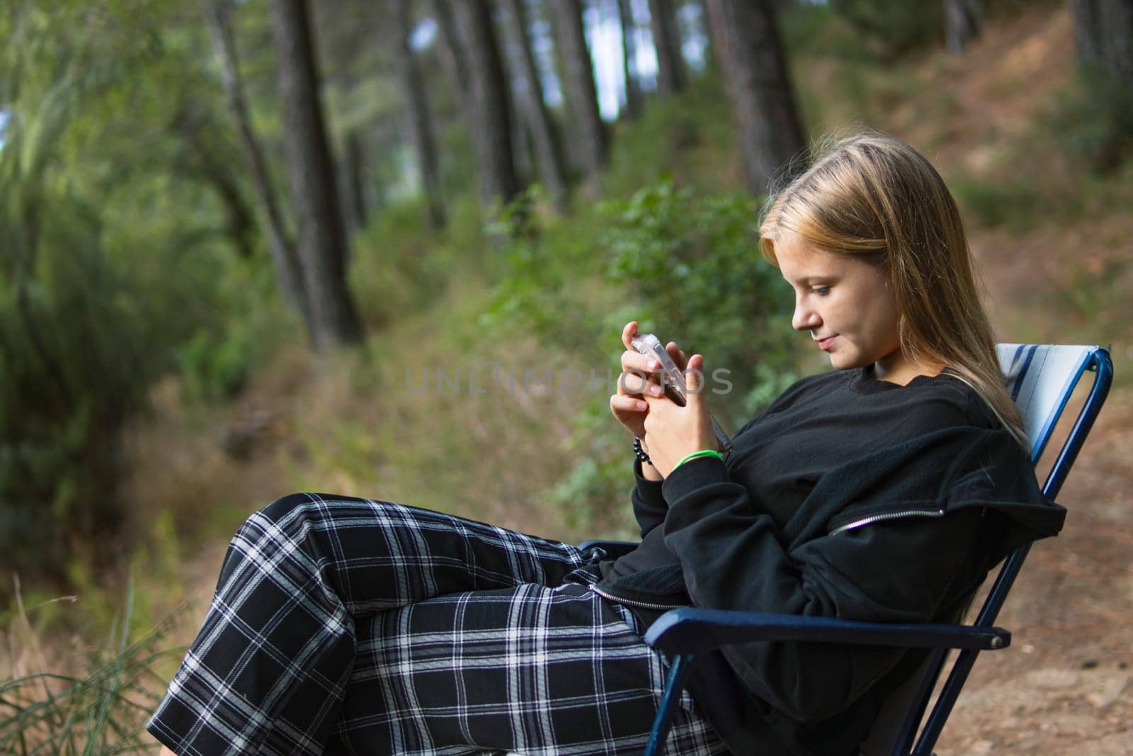 a teenage girl in casual clothes sits on a chair in a Camping and looks at the phone, by PopOff