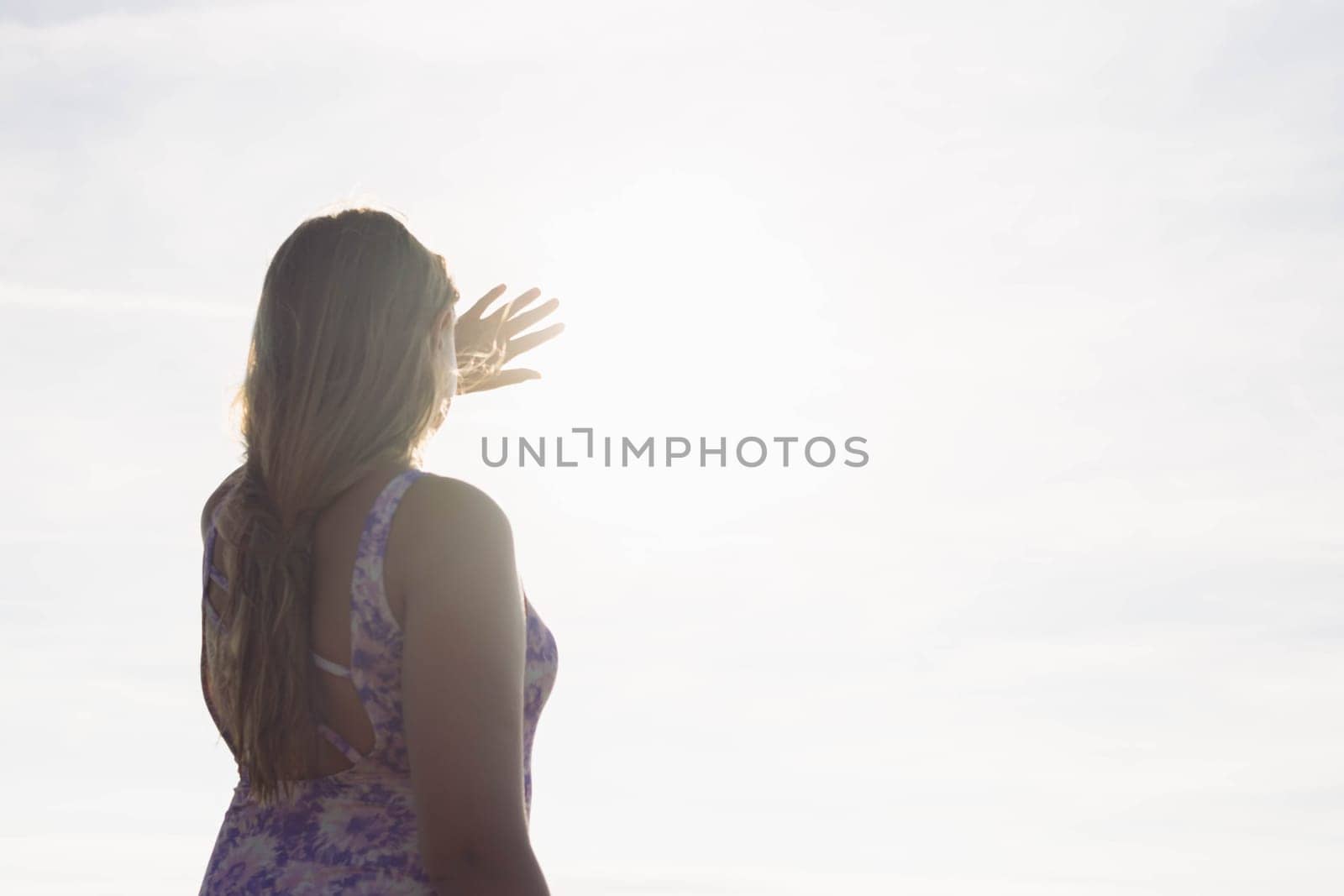 girl with long blond hair stands facing the sunset and the ocean in a bathing suit and covers the sun with her hand, there is a place for an inscription . Beautiful background. High quality photo