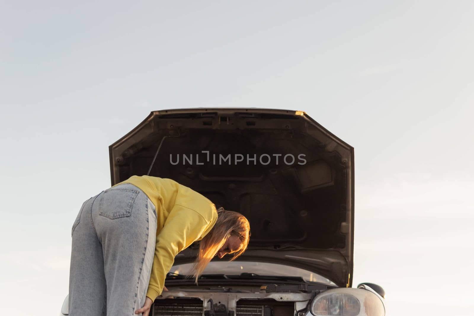 a girl with blond hair in a yellow sweater and jeans, stands with the hood of the car open and looks at what has broken, bent over to the hood on the road. photo angle from the bottom by PopOff