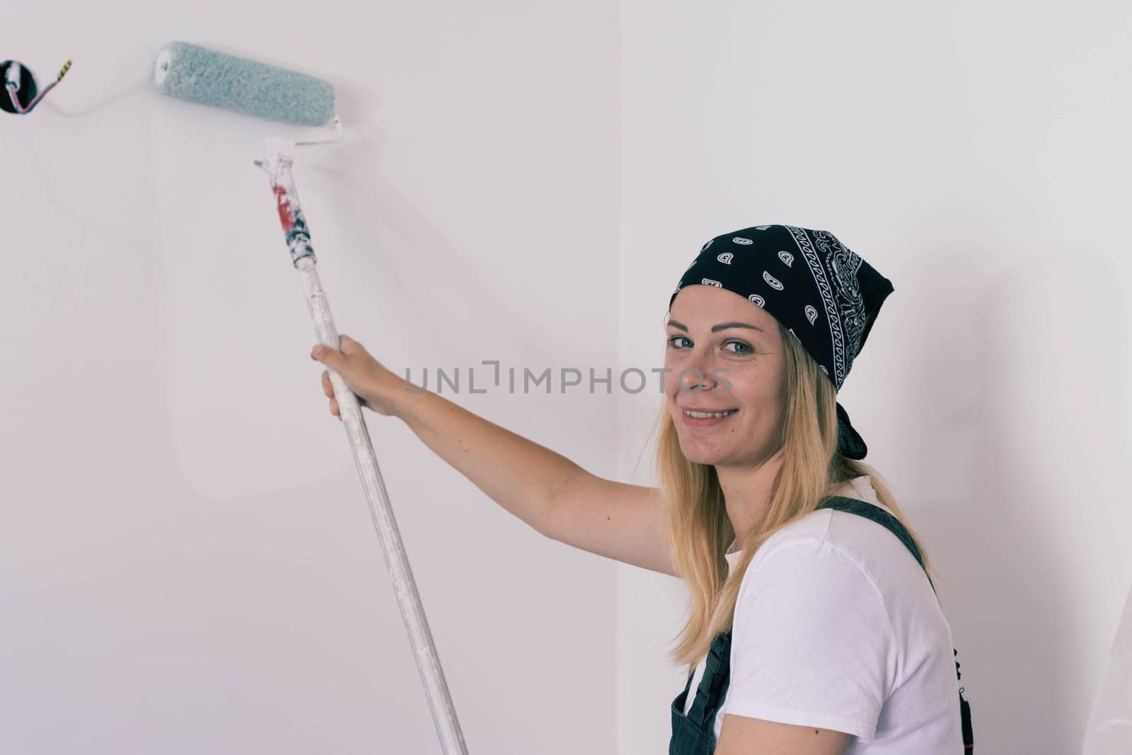 a happy girl of European appearance with a black bandana on her head is dressed in a white T-shirt and a dark work overall, paints a white wall with a roller. Gender equality High quality photo