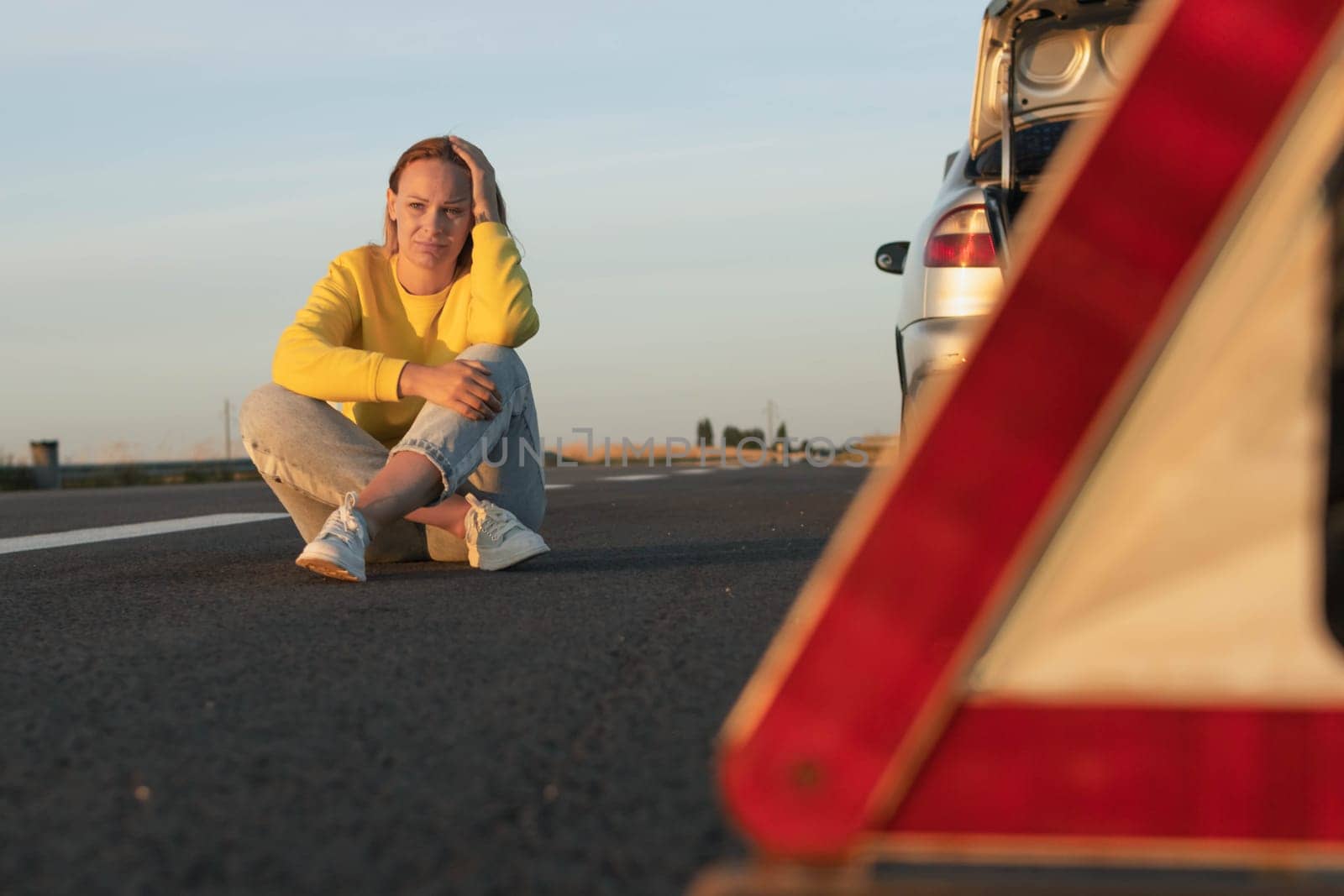 a girl sits on the pavement near a broken car, upset, worries next to it is a white-red emergency stop sign. The girl is waiting for help with a broken car, there is a place for an inscription by PopOff