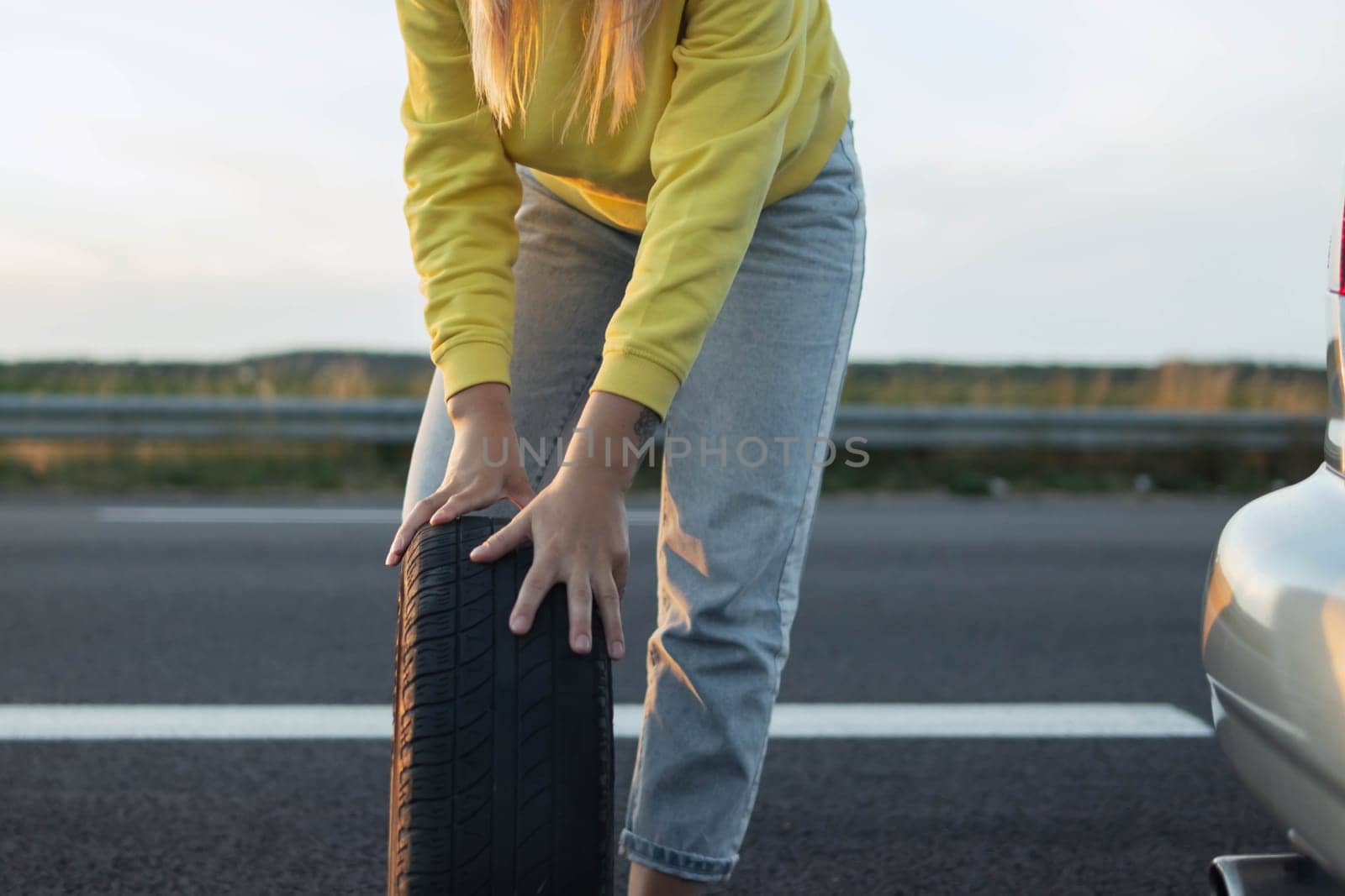 a girl in casual clothes stands near the rear wheel of the car and rolls a spare wheel to change it.the wheel in front is a temporary stop sign blurred place for an inscription. High quality photo