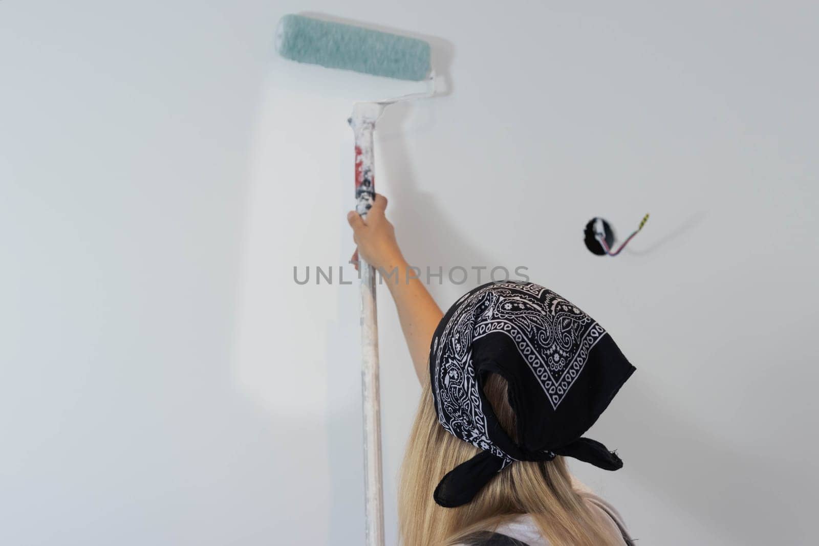 a girl with blond hair in a dark bandana, paints the wall with a roller in white at home. stands with her back to the camera. there is a place for milking the inscription. High quality photo