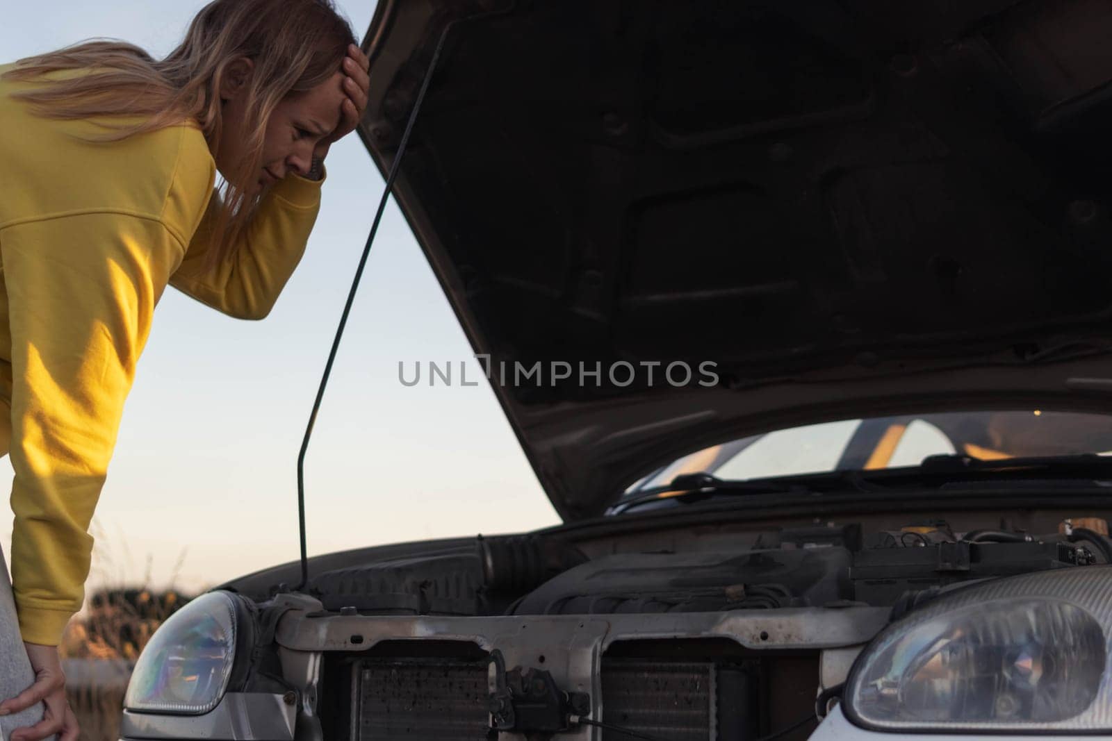 a girl with blond hair in a yellow sweater and jeans, stands with the hood of the car open and looks with a surprised look at what has broken holding her head with her hand A car breakdown on the road by PopOff