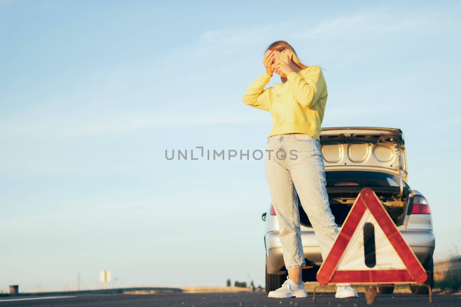 woman stands near a broken car with an open trunk, speaks on the phone, holds her head, worries next to a white-red emergency stop sign.on the left there is a place for an inscription by PopOff