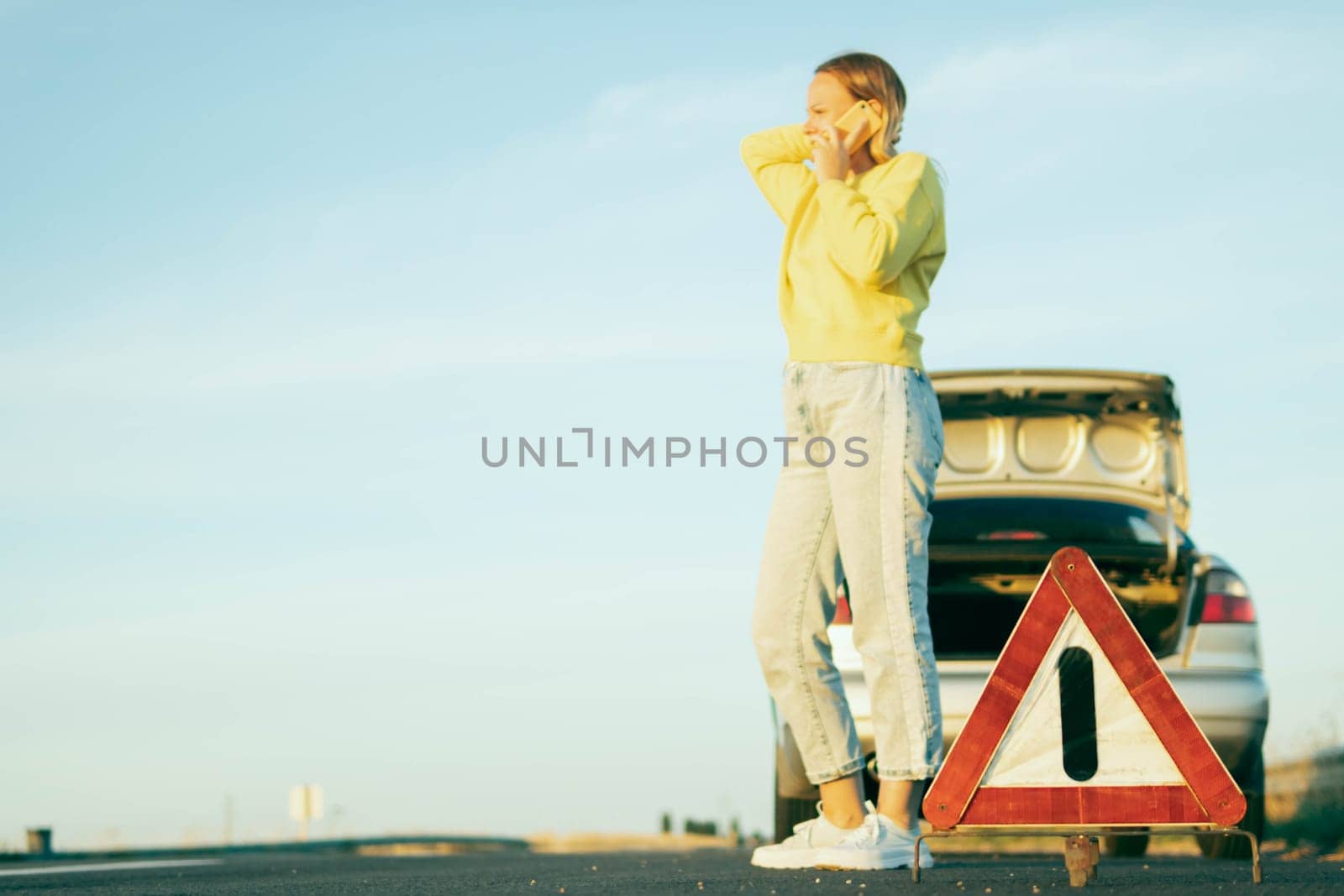 a woman in casual clothes of European appearance stands near a broken car with an open trunk, speaks on the phone, she is worried nearby there is a white-red emergency stop sign.High quality photo