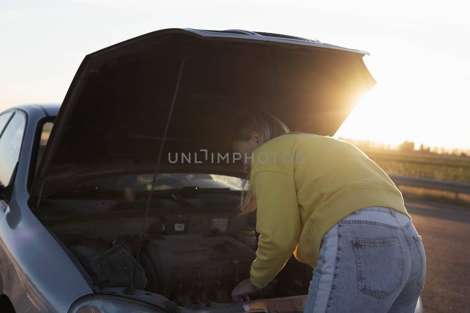 a girl with blond hair in casual clothes stands with an open hood at sunset and looks at what has broken. A car breakdown on the road. There is a place for an inscription on the left by PopOff