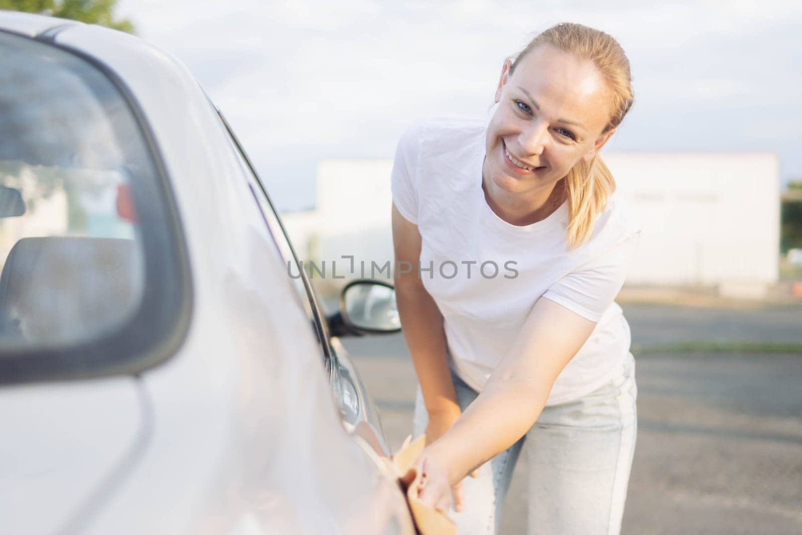 a cheerful girl with blond hair in a white T-shirt and jeans, bending over wipes the car after washing with a special yellow rag with microfiber. Looks at the camera. High quality photo
