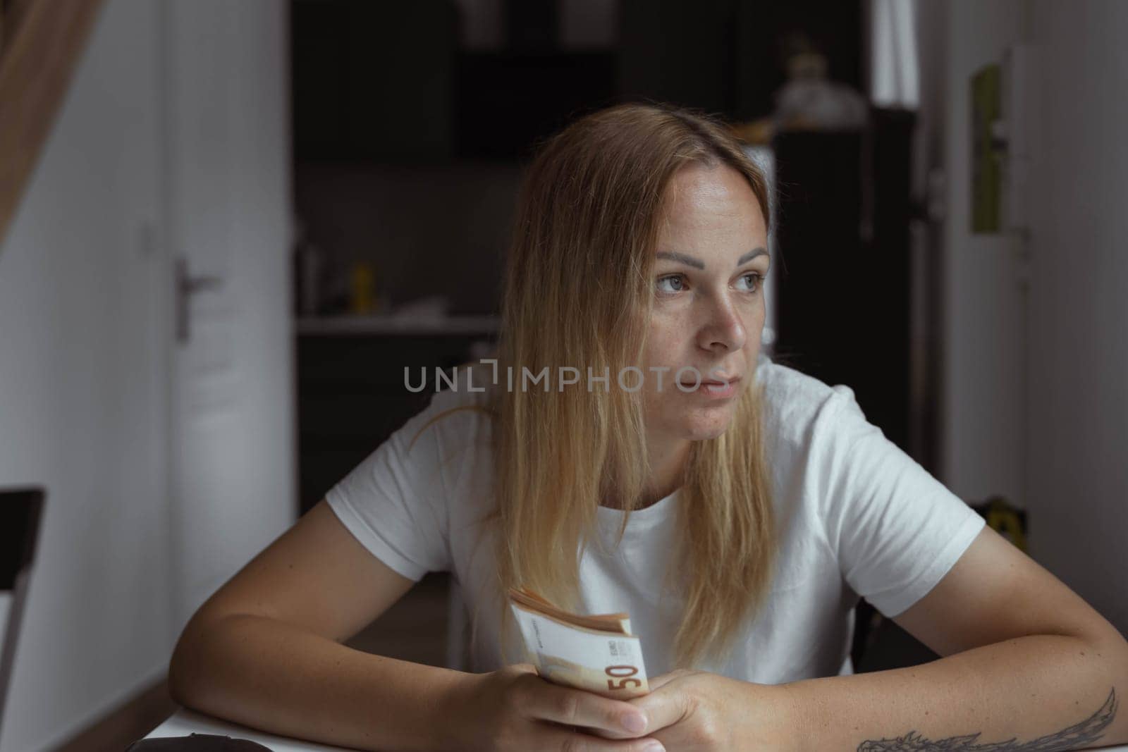 a girl sits indoors at a white table with her hands folded and the table is thoughtful looks to the side on the girl left hand has a tattoo, in her hands the girl holds euros of 50 salaries. by PopOff