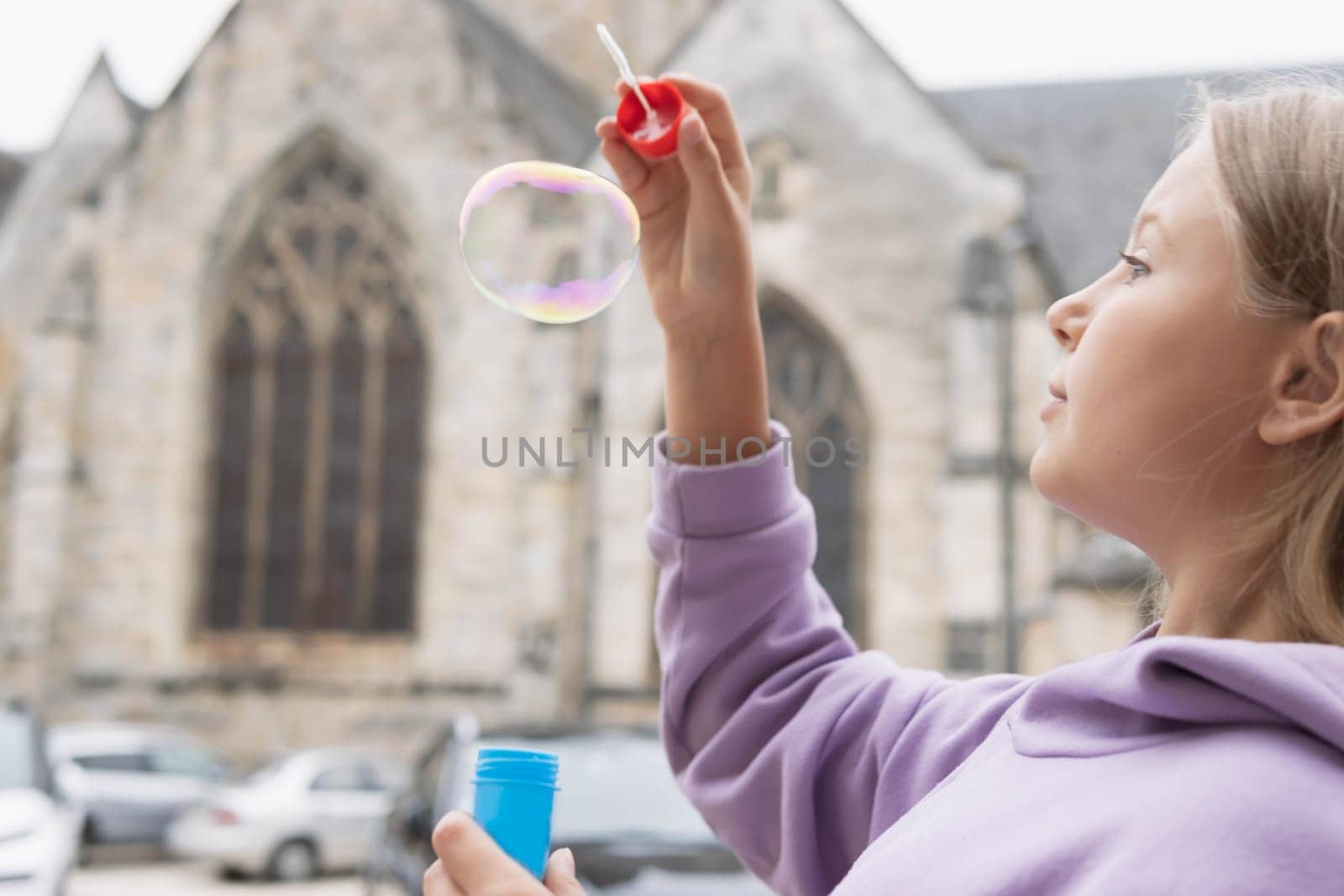 a girl of European appearance with blond hair in a lilac sweater plays with bubbles on the street. High quality photo