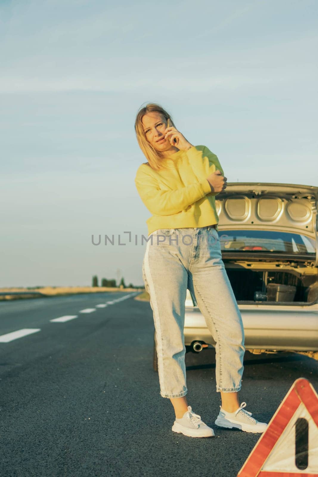 a girl in a yellow sweater and jeans stands on the road near a broken car and speaks on the phone upset, help peeps out in the foreground an emergency stop sign girl's car broke down on the road by PopOff