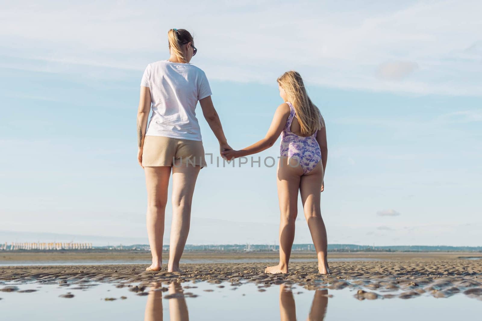 mothers in a white t-shirt and brown shorts with a daughter in a purple swimsuit walk along the beach in full growth photo.Beautiful landscape of the beach by PopOff