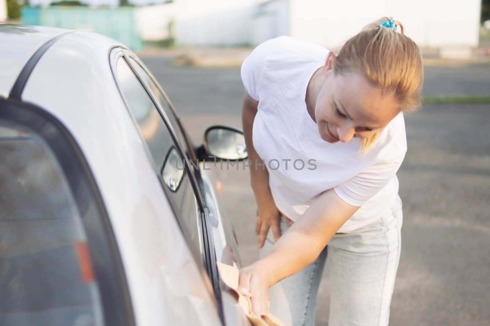 a girl of European appearance with blond hair in a white T-shirt and jeans, bending over wipes the car after washing with a special yellow rag with microfiber. The concept of cleanliness by PopOff