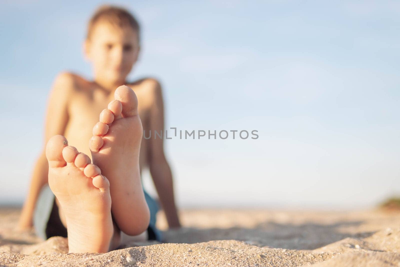 a child of European appearance with blond hair sits on the sand on the beach close-up of legs and feet in focus the body of a blurry boy on the right there is a place for an inscription by PopOff