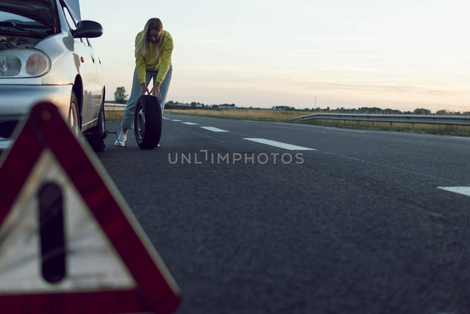 girl in stands near the rear wheel of the car and rolls a spare wheel to change it.A car breakdown, the wheel in front is a temporary stop sign blurred on the right there is a place for an inscription by PopOff