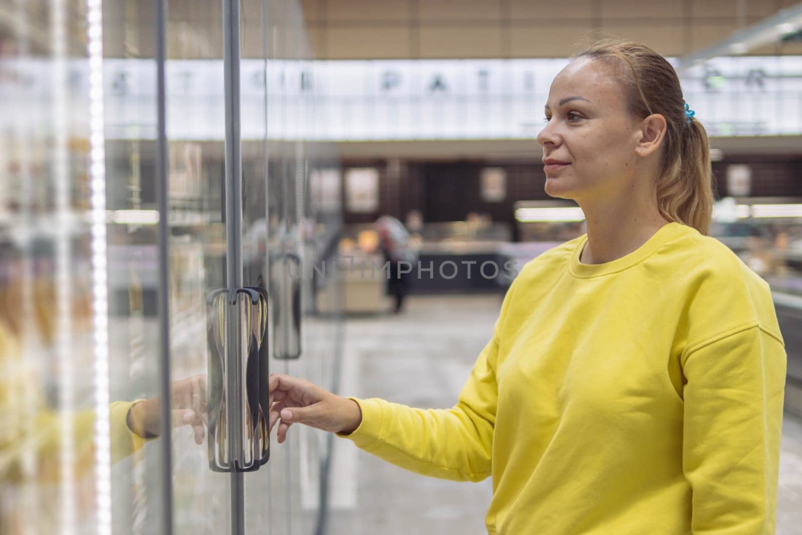 a cheerful girl of European appearance with blond hair tied in a ponytail with a blue elastic band in a yellow jacket, chooses products in a refrigerated display case in a grocery store by PopOff