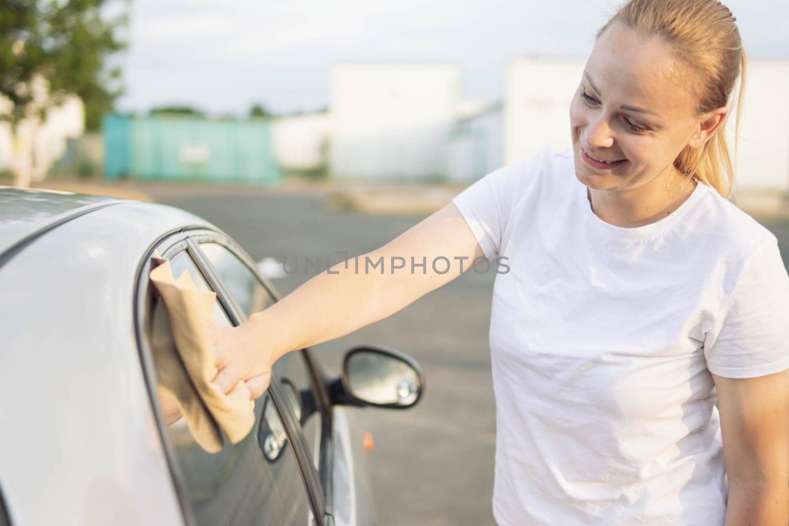 smiling girl of European appearance with blond hair tied in a ponytail in a white T-shirt, wipes the side windows in the car with a special yellow rag while milking the windows. High quality photo