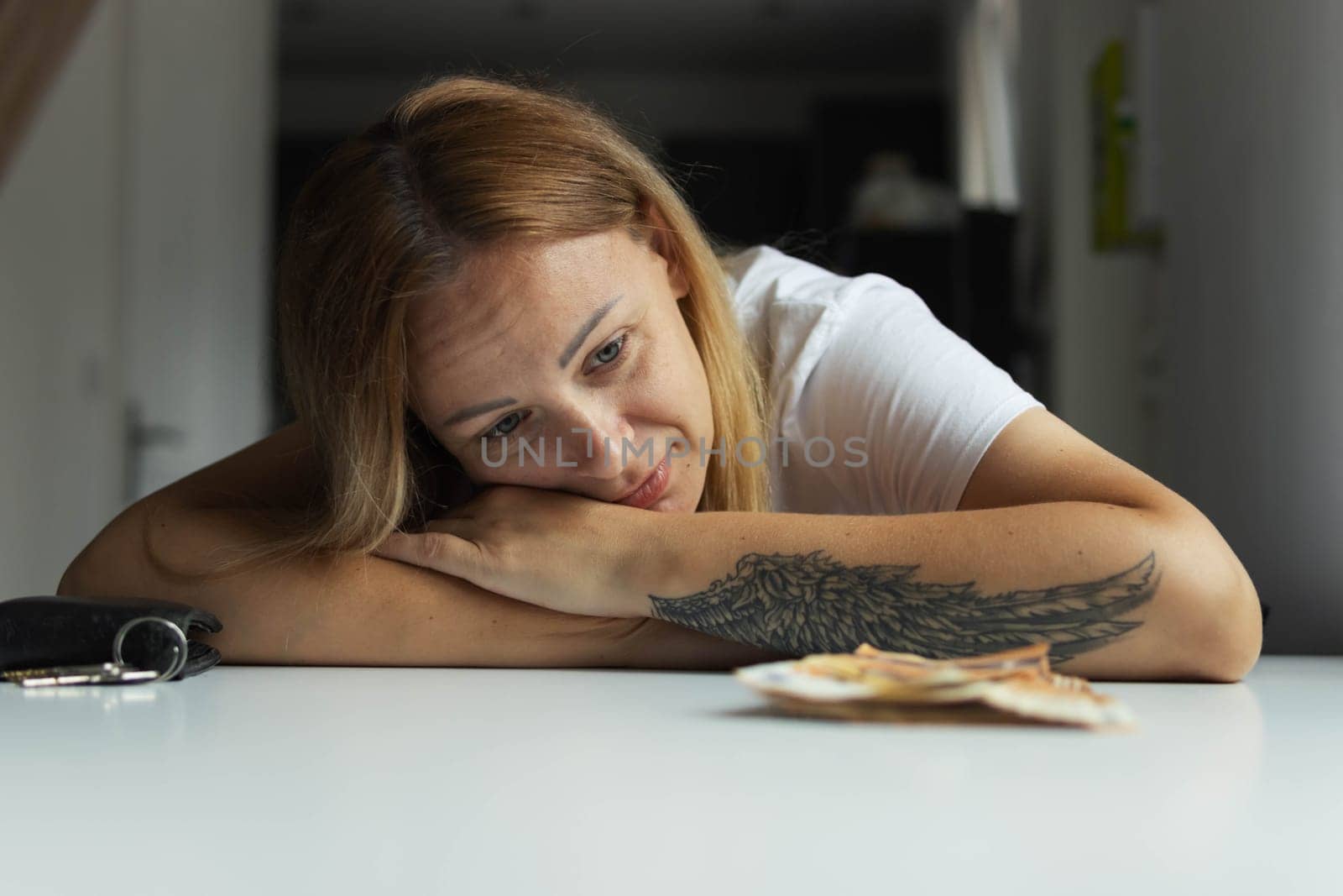 a girl of European appearance sits indoors at a white table with her head on her hands and the table is sad on the left hand of the girl with a tattoo, money lies nearby. High quality photo