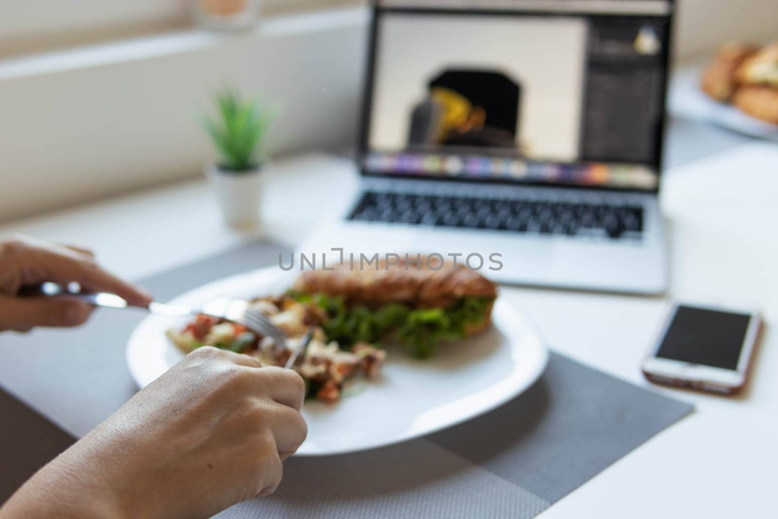 a girl sits at a white table and has breakfast, focus on hands close-up blurred background, next to it is a laptop, phone, indoor flower, there is a place for an inscription. High quality photo