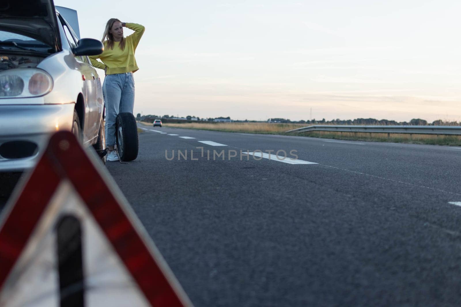 a girl in casual clothes stands near the rear wheel of the car and rolls a spare wheel to change it.the wheel in front is a temporary stop sign blurred place for an inscription. High quality photo