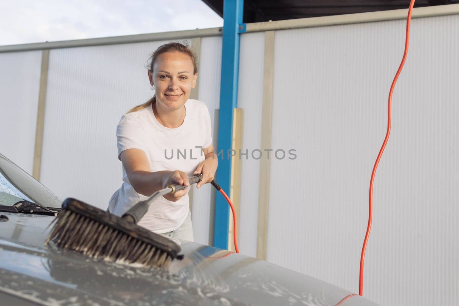 a cheerful girl in a white t-shirt with blond hair tied in a ponytail washes a gray car with a brush at the sink.The concept of work at the sink by PopOff