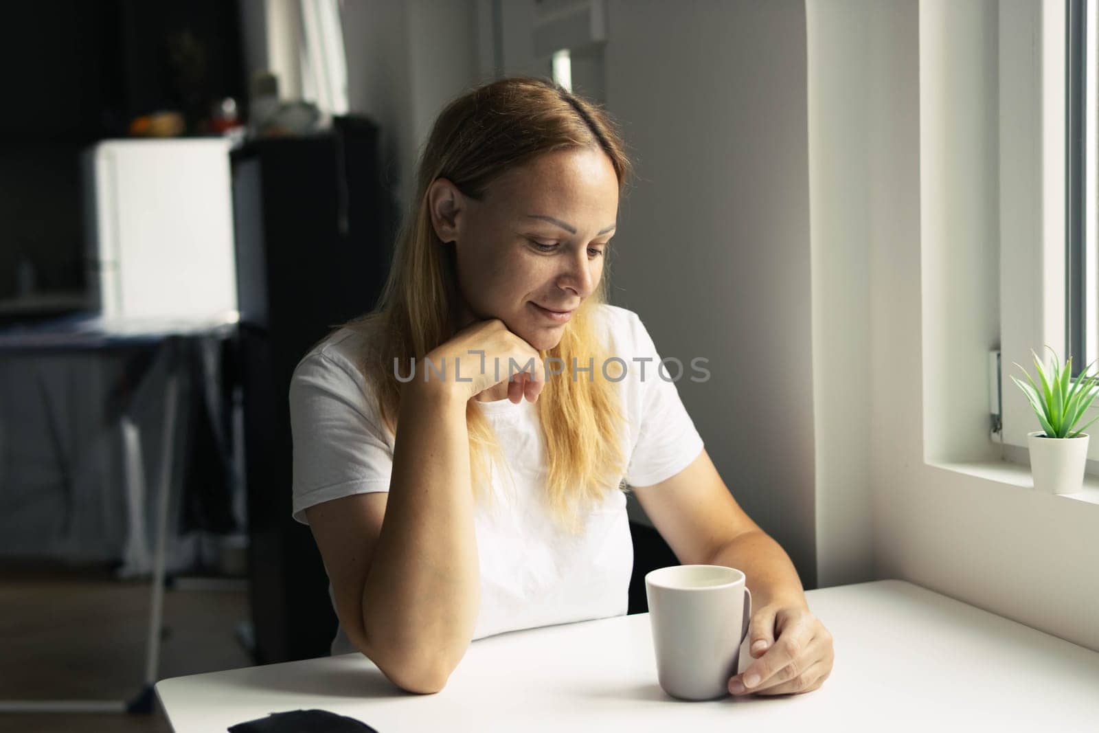 a girl of European appearance in a white T-shirt sits at a white table at home in the kitchen in the morning drinks coffee or tea from a light cup and daydreams, looks at the cup. High quality photo